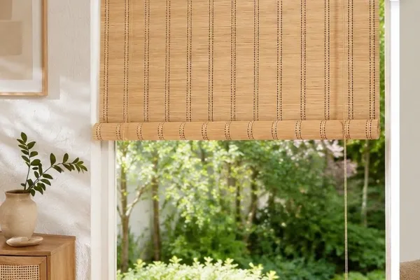 Natural bamboo chick blinds half-rolled over a tall window with a garden view, in a soft modern Indian living room with cane cabinet and plants.