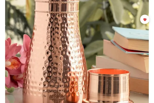 Hammered copper water bottle with cup-style lid on a bedside table next to a lotus flower and a stack of books.